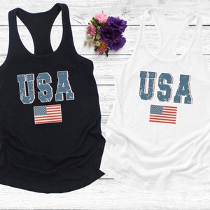 K&ouml;nnte beinhalten: Zwei Tanktops mit einem abgenutzten USA-Motiv und einer kleinen amerikanischen Flagge. Das Tanktop links ist marineblau und das Tanktop rechts ist wei&szlig;.