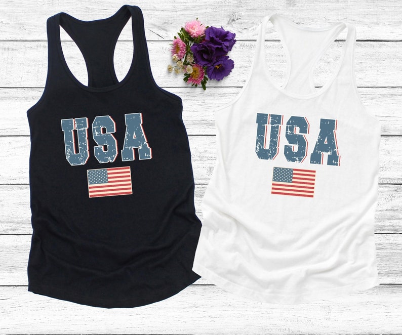 K&ouml;nnte beinhalten: Zwei Tanktops mit einem abgenutzten USA-Motiv und einer kleinen amerikanischen Flagge. Das Tanktop links ist marineblau und das Tanktop rechts ist wei&szlig;.