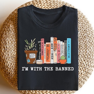 Może przedstawiać: Czarny t-shirt z kolorową grafiką przedstawiającą rząd książek, roślinę doniczkową i mały kaktus. Pod książkami wydrukowano napis "I'M WITH THE BANNED". Projekt umieszczono na okrągłym, plecionym tle.