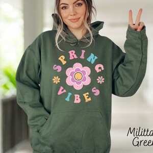 Op de afbeelding: Een groene hoodie met een witte en roze grafische afbeelding met de tekst "Spring Vibes" en een bloemendesign.