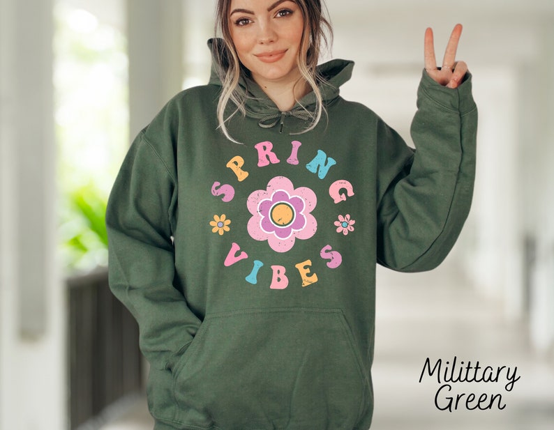 Op de afbeelding: Een groene hoodie met een witte en roze grafische afbeelding met de tekst "Spring Vibes" en een bloemendesign.
