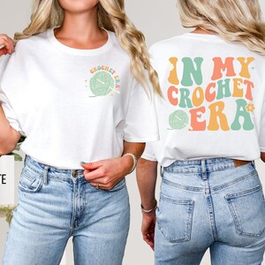 K&ouml;nnte beinhalten: Wei&szlig;es T-Shirt mit dem Text "CROCHET ERA" und einer Garnkn&auml;uel-Grafik auf der Vorderseite. Auf der R&uuml;ckseite des Shirts steht der Text "IN MY CROCHET ERA" in Retro-Farben. Ein lustiges, l&auml;ssiges Oberteil f&uuml;r Handwerker.