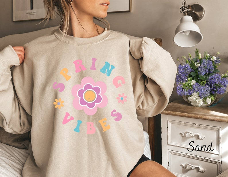 Op de afbeelding: Een beige sweatshirt met een kleurrijk grafisch ontwerp met de tekst "Spring Vibes" en een bloem in het midden.