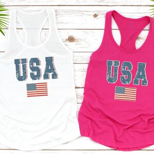 K&ouml;nnte beinhalten: Zwei Tanktops mit dem Wort "USA" in einer verwitterten blauen Schrift darauf gedruckt. Die Oberteile sind wei&szlig; und rosa und jedes hat eine kleine amerikanische Flagge unter dem Wort "USA" gedruckt.