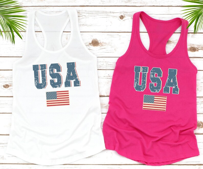 K&ouml;nnte beinhalten: Zwei Tanktops mit dem Wort "USA" in einer verwitterten blauen Schrift darauf gedruckt. Die Oberteile sind wei&szlig; und rosa und jedes hat eine kleine amerikanische Flagge unter dem Wort "USA" gedruckt.