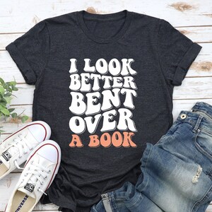 以下が含まれることがあります： 暗いグレーのTシャツに「I look better bent over a book」という文字が白と桃色の文字で書かれています。文字はレトロなフォントスタイルです。