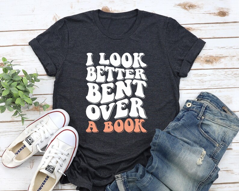以下が含まれることがあります： 暗いグレーのTシャツに「I look better bent over a book」という文字が白と桃色の文字で書かれています。文字はレトロなフォントスタイルです。