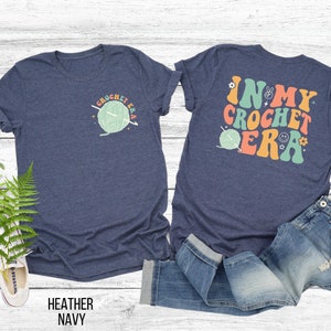 K&ouml;nnte beinhalten: Marineblaues T-Shirt mit einem H&auml;kel-Design. Auf der Vorderseite befindet sich eine kleine Grafik mit dem Text "CROCHET ERA". Auf der R&uuml;ckseite des Shirts steht der Satz "IN MY CROCHET ERA" in farbenfrohen Buchstaben im Retro-Stil.