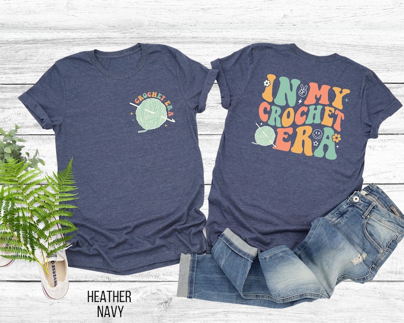 K&ouml;nnte beinhalten: Marineblaues T-Shirt mit einem H&auml;kel-Design. Auf der Vorderseite befindet sich eine kleine Grafik mit dem Text "CROCHET ERA". Auf der R&uuml;ckseite des Shirts steht der Satz "IN MY CROCHET ERA" in farbenfrohen Buchstaben im Retro-Stil.