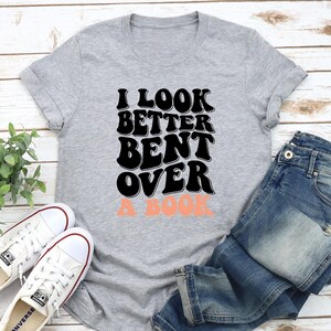 以下が含まれることがあります： グレーのTシャツに「I look better bent over a book」という文字が黒と桃色で書かれています。