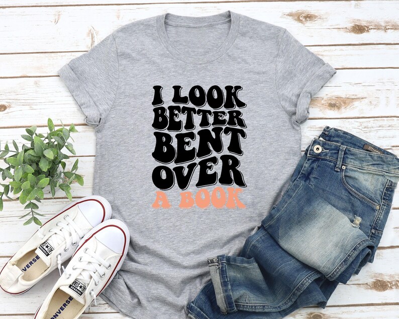 以下が含まれることがあります： グレーのTシャツに「I look better bent over a book」という文字が黒と桃色で書かれています。