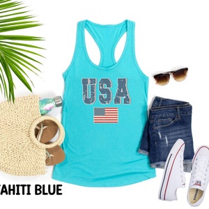 K&ouml;nnte beinhalten: Ein t&uuml;rkisfarbenes Tanktop mit einem verwaschenen blauen "USA"-Aufdruck und einer kleinen amerikanischen Flagge. Das Tanktop liegt auf einem wei&szlig;en Hintergrund mit einer Strohtasche, Sonnenbrille, Jeans-Shorts und wei&szlig;en Turnschuhen.