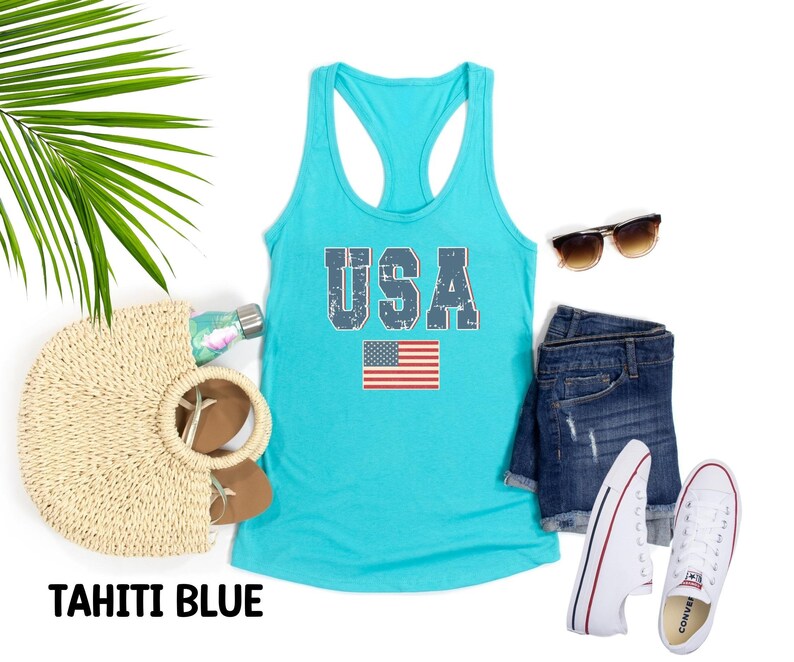 K&ouml;nnte beinhalten: Ein t&uuml;rkisfarbenes Tanktop mit einem verwaschenen blauen "USA"-Aufdruck und einer kleinen amerikanischen Flagge. Das Tanktop liegt auf einem wei&szlig;en Hintergrund mit einer Strohtasche, Sonnenbrille, Jeans-Shorts und wei&szlig;en Turnschuhen.