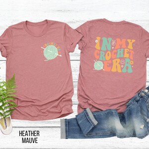 K&ouml;nnte beinhalten: Heather-Mauvefarbenes T-Shirt mit einem Front-Grafik einer Garnkugel und dem Text "CROCHET LIFE". Auf der R&uuml;ckseite des Shirts steht der Text "IN MY CROCHET ERA" mit einem Friedenszeichen, einem Smiley und einer Garnkugel-Grafik.