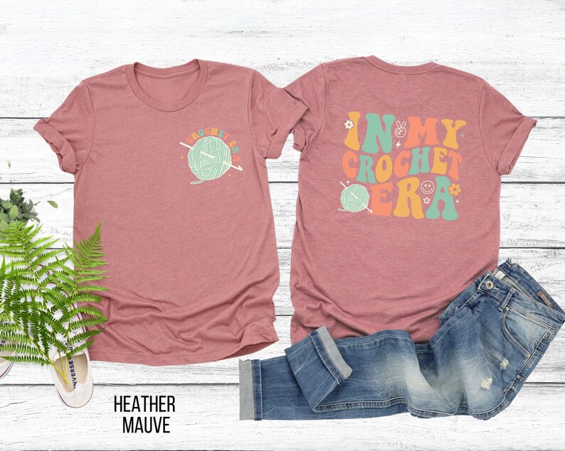K&ouml;nnte beinhalten: Heather-Mauvefarbenes T-Shirt mit einem Front-Grafik einer Garnkugel und dem Text "CROCHET LIFE". Auf der R&uuml;ckseite des Shirts steht der Text "IN MY CROCHET ERA" mit einem Friedenszeichen, einem Smiley und einer Garnkugel-Grafik.
