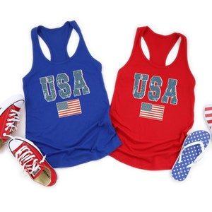 K&ouml;nnte beinhalten: Zwei Tanktops mit dem Wort "USA" darauf gedruckt in einer verwitterten Schriftart. Die Tanktops sind rot und blau und zeigen eine kleine amerikanische Flagge.