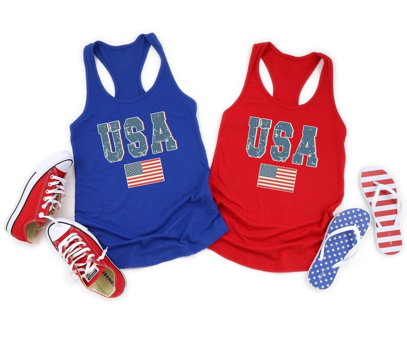K&ouml;nnte beinhalten: Zwei Tanktops mit dem Wort "USA" darauf gedruckt in einer verwitterten Schriftart. Die Tanktops sind rot und blau und zeigen eine kleine amerikanische Flagge.
