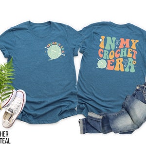 K&ouml;nnte beinhalten: Ein teal-farbenes T-Shirt mit dem Text "CROCHET ERA" und einer Garnkn&auml;uel-Grafik auf der Vorderseite. Auf der R&uuml;ckseite des Shirts steht der Text "IN MY CROCHET ERA" in Retro-Farben. Das Shirt wird mit einer blauen Jeans und wei&szlig;en Turnschuhen kombiniert.