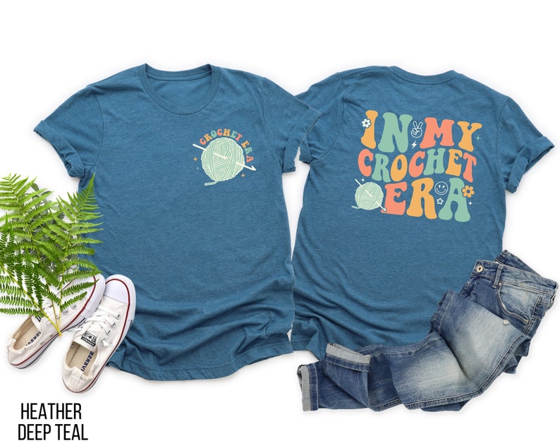 K&ouml;nnte beinhalten: Ein teal-farbenes T-Shirt mit dem Text "CROCHET ERA" und einer Garnkn&auml;uel-Grafik auf der Vorderseite. Auf der R&uuml;ckseite des Shirts steht der Text "IN MY CROCHET ERA" in Retro-Farben. Das Shirt wird mit einer blauen Jeans und wei&szlig;en Turnschuhen kombiniert.