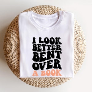 以下が含まれることがあります： 白いTシャツに黒い文字で「本に身を乗り出して見た方が良い」と書かれています。テキストはレトロなフォントスタイルです。「本」という単語はピーチカラーです。