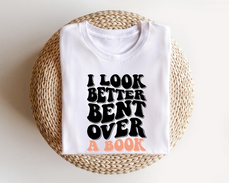 以下が含まれることがあります： 白いTシャツに黒い文字で「本に身を乗り出して見た方が良い」と書かれています。テキストはレトロなフォントスタイルです。「本」という単語はピーチカラーです。