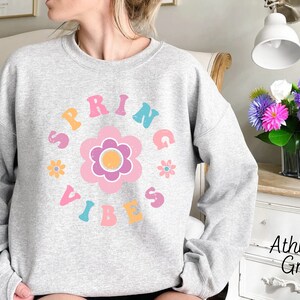 Op de afbeelding: Een grijs sweatshirt met een kleurrijk grafisch ontwerp met de tekst "Spring Vibes" in een retro lettertype. De grafiek bevat een grote roze bloem met een gele kern en kleinere roze bloemen eromheen.