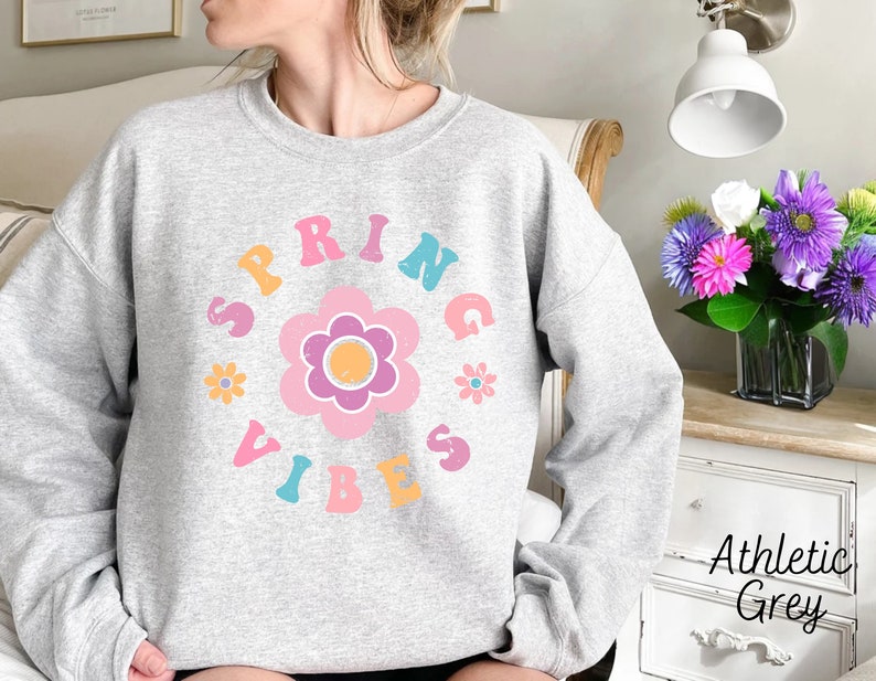 Op de afbeelding: Een grijs sweatshirt met een kleurrijk grafisch ontwerp met de tekst "Spring Vibes" in een retro lettertype. De grafiek bevat een grote roze bloem met een gele kern en kleinere roze bloemen eromheen.
