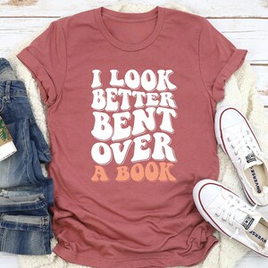 以下が含まれることがあります： 文字が桃色の背景になっている、白文字で「I look better bent over a book」と書かれた、マルーンのTシャツです。