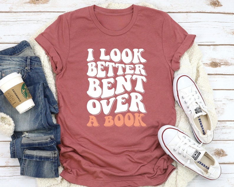 以下が含まれることがあります： 文字が桃色の背景になっている、白文字で「I look better bent over a book」と書かれた、マルーンのTシャツです。