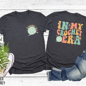 K&ouml;nnte beinhalten: Dunkelgraue Heather-T-Shirts mit H&auml;kelmotiven. Ein Shirt zeigt eine kleine Grafik von Garn und Nadeln mit dem Text "I Crochet Era". Das andere Shirt hat den Text "In My Crochet Era" mit einer Garnkn&auml;uel-Grafik.