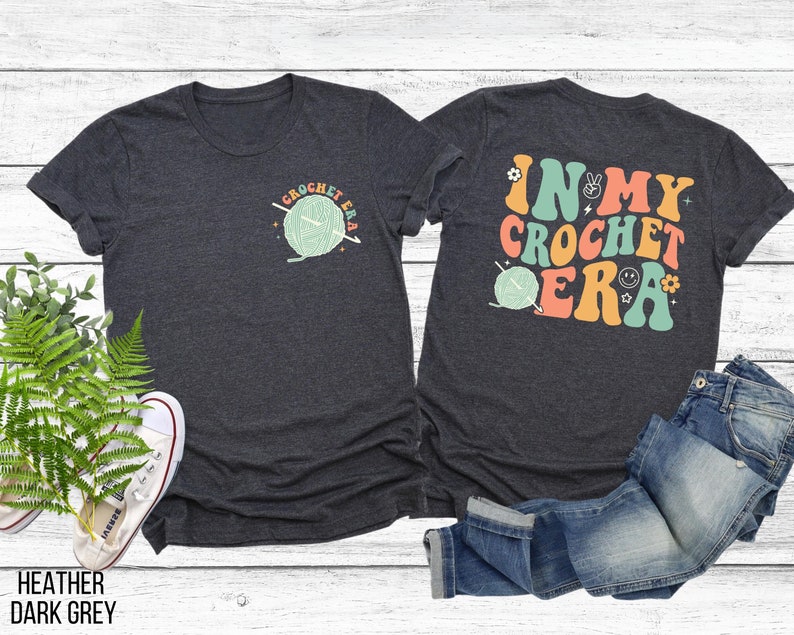 K&ouml;nnte beinhalten: Dunkelgraue Heather-T-Shirts mit H&auml;kelmotiven. Ein Shirt zeigt eine kleine Grafik von Garn und Nadeln mit dem Text "I Crochet Era". Das andere Shirt hat den Text "In My Crochet Era" mit einer Garnkn&auml;uel-Grafik.
