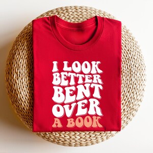 以下が含まれることがあります： 赤いTシャツに白い文字で「私は本に身を乗り出して見た方が良い」と書かれています。文字はレトロなフォントスタイルです。