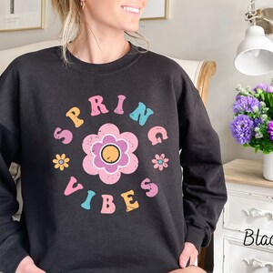 Op de afbeelding: Zwart sweatshirt met een grafische print van een bloem en de tekst "SPRING VIBES" in kleurrijke letters.