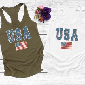 K&ouml;nnte beinhalten: Zwei Tanktops mit dem Wort "USA" in blau bedruckt, das einen abgenutzten Look hat. Das Wort ist mittig auf den Tanktops platziert, darunter ist eine kleine amerikanische Flagge gedruckt. Ein Tanktop ist olivgr&uuml;n und das andere ist wei&szlig;.
