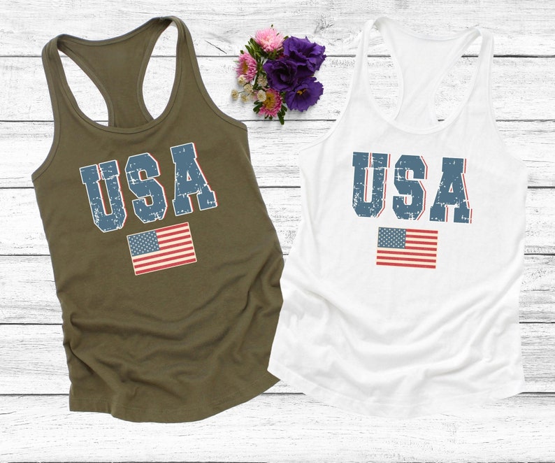K&ouml;nnte beinhalten: Zwei Tanktops mit dem Wort "USA" in blau bedruckt, das einen abgenutzten Look hat. Das Wort ist mittig auf den Tanktops platziert, darunter ist eine kleine amerikanische Flagge gedruckt. Ein Tanktop ist olivgr&uuml;n und das andere ist wei&szlig;.