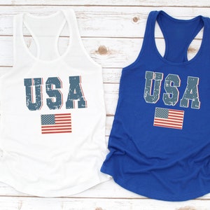 K&ouml;nnte beinhalten: Zwei Tanktops mit dem Wort "USA" in blau bedruckt, das einen abgenutzten Look hat. Die Tanktops sind wei&szlig; und blau. Das wei&szlig;e Tanktop hat eine rot-wei&szlig;-blaue amerikanische Flagge unter dem Wort "USA" gedruckt. Das blaue Tanktop hat eine rot-wei&szlig;-blaue amerikanische Flagge rechts neben dem Wort "USA" gedruckt.