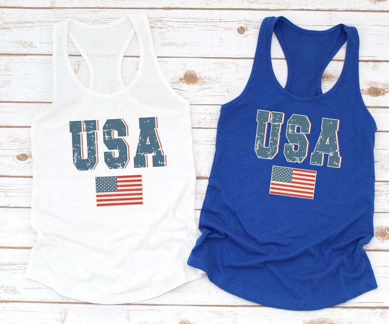 K&ouml;nnte beinhalten: Zwei Tanktops mit dem Wort "USA" in blau bedruckt, das einen abgenutzten Look hat. Die Tanktops sind wei&szlig; und blau. Das wei&szlig;e Tanktop hat eine rot-wei&szlig;-blaue amerikanische Flagge unter dem Wort "USA" gedruckt. Das blaue Tanktop hat eine rot-wei&szlig;-blaue amerikanische Flagge rechts neben dem Wort "USA" gedruckt.