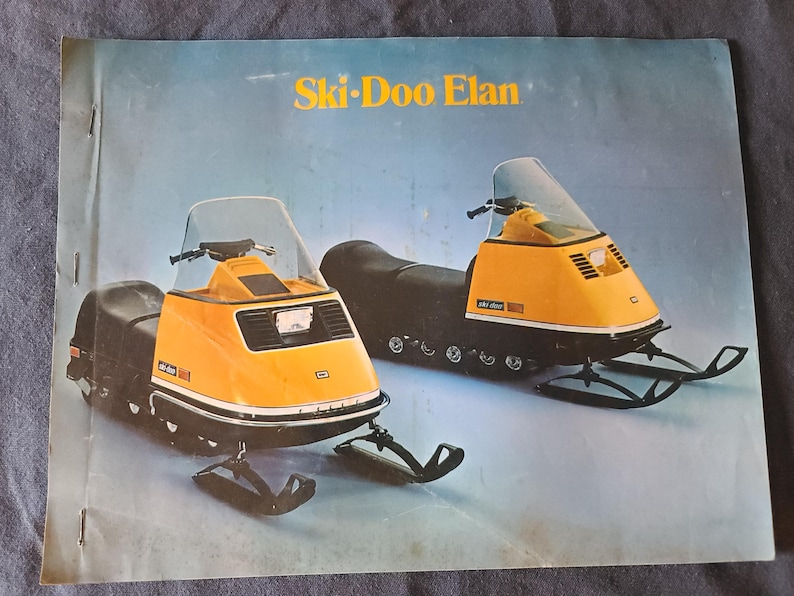 May include: A vintage brochure showcasing two yellow and black Ski-Doo Elan snowmobiles. The snowmobiles have a distinctive design with a windshield, black seats, and skis. The brochure has the words "Ski-Doo Elan" in gold lettering.