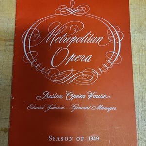 May include: A vintage orange program cover with white script. The text reads "Metropolitan Opera" in an ornate font, with "Boston Opera House" and "Season of 1949" below. The cover is slightly worn.