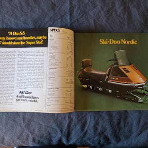 May include: A vintage magazine spread showcasing a Ski-Doo snowmobile advertisement. The right page features a brown and black snowmobile with the text "Ski-Doo Nordic" on a green background. The left page has text that reads "'74 Elan S/S" and "A million machines can teach you a lot."