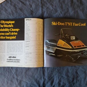 May include: A vintage magazine spread advertising a Ski-Doo snowmobile. The snowmobile is black, yellow, and orange. Text includes "'74 Olympique" and "Ski-Doo T'NT Fan Cool". The magazine is open on a dark blue surface.