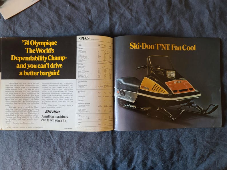 May include: A vintage magazine spread advertising a Ski-Doo snowmobile. The snowmobile is black, yellow, and orange. Text includes "'74 Olympique" and "Ski-Doo T'NT Fan Cool". The magazine is open on a dark blue surface.