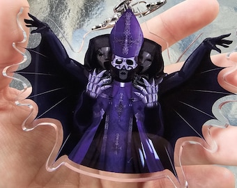 May include: A clear acrylic keychain featuring a purple and black illustration of a skeletal figure in a bishop's hat and robes, with bat-like wings. The keychain has a silver-colored metal clasp.