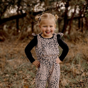 May include: A young girl wearing a black long-sleeved shirt and a pink and black leopard print jumpsuit with ruffles on the shoulders. She is standing in a grassy area with fallen leaves.