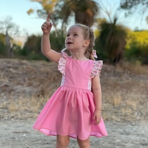 May include: A young girl wearing a pink dress with floral print accents and white ruffle trim. She is wearing gold sandals and pointing upwards.