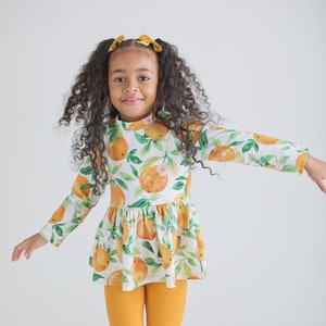 Puede incluir: Una niña lleva una camiseta blanca de manga larga con un estampado de naranjas y hojas, combinada con leggings color mostaza. También lleva un lazo color mostaza en el pelo y zapatos marrones.