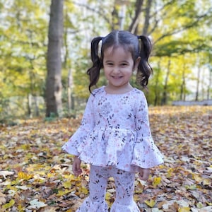 May include: A young girl wearing a white and pink floral print top and matching bell bottom pants. She is standing in a pile of autumn leaves.