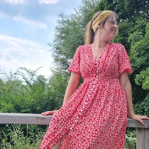 May include: A red floral print dress with short sleeves and a button-down front. The dress has a V-neckline and a slit on the side. The dress is knee-length and is worn with a yellow headband.