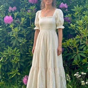 May include: A floor-length cream-colored dress with a square neckline, short puff sleeves with ruffled edges, and a tiered skirt. The dress features a fitted bodice and a long, flowing silhouette that reaches the ankles, paired with brown sandals.