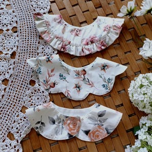May include: Three white fabric collars with different floral patterns. The top collar has a pink and white floral pattern with ruffled edges. The middle collar has a white background with a blue and green floral pattern. The bottom collar has a white background with a peach and gray floral pattern.